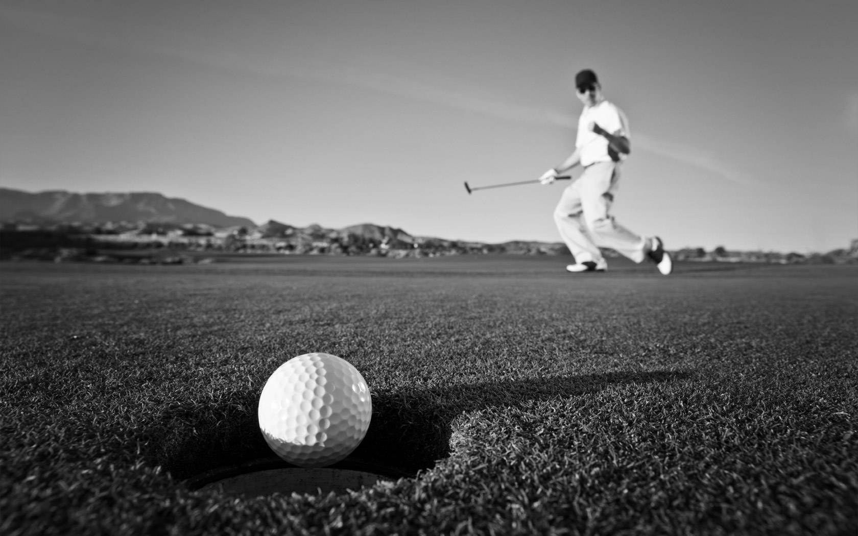 Czarno-białe zdjęcie: piłka golfowa na skraju dołka, w tle golfista